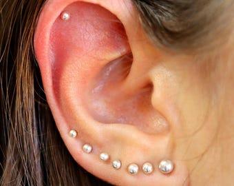 Può includere: Primo piano di un orecchio con più orecchini a perno d'argento. Gli orecchini sono disposti in linea, partendo dalla parte superiore dell'orecchio e scendendo fino al lobo. Gli orecchini sono di diverse dimensioni, con il più grande in basso.