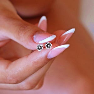 May include: A pair of silver flower stud earrings with a black center. The earrings are shown on a person's hand with white nail polish.