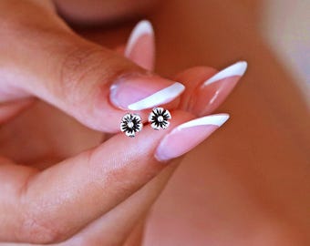 May include: A pair of silver flower stud earrings with a black center. The earrings are shown on a person's hand with white nail polish.