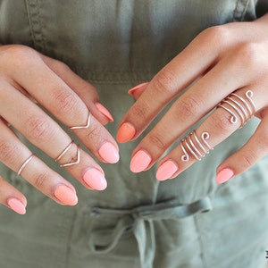 Pode incluir: Uma mão com vários anéis de prata, incluindo um anel espiral e um anel com um design em forma de V. As unhas estão pintadas de uma cor pêssego claro.