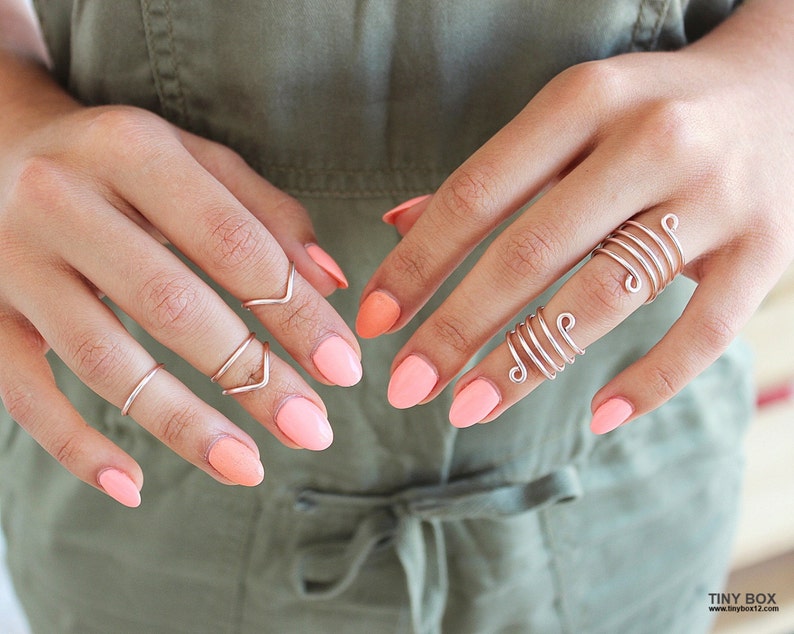 May include: A hand wearing several silver rings, including a spiral ring and a ring with a V-shaped design. The nails are painted a light peach color.