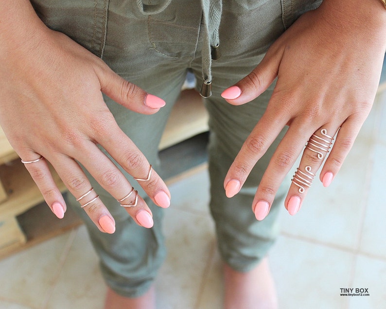 May include: Close-up of hands wearing multiple silver rings on several fingers. The rings are thin and delicate, with some featuring a simple spiral design. The nails are painted a light pink color.