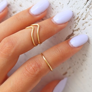Pode incluir: Um conjunto de três anéis de ouro em um dedo. Os anéis são finos e delicados, com um design simples. O anel no dedo do meio tem a forma de um duplo chevron.