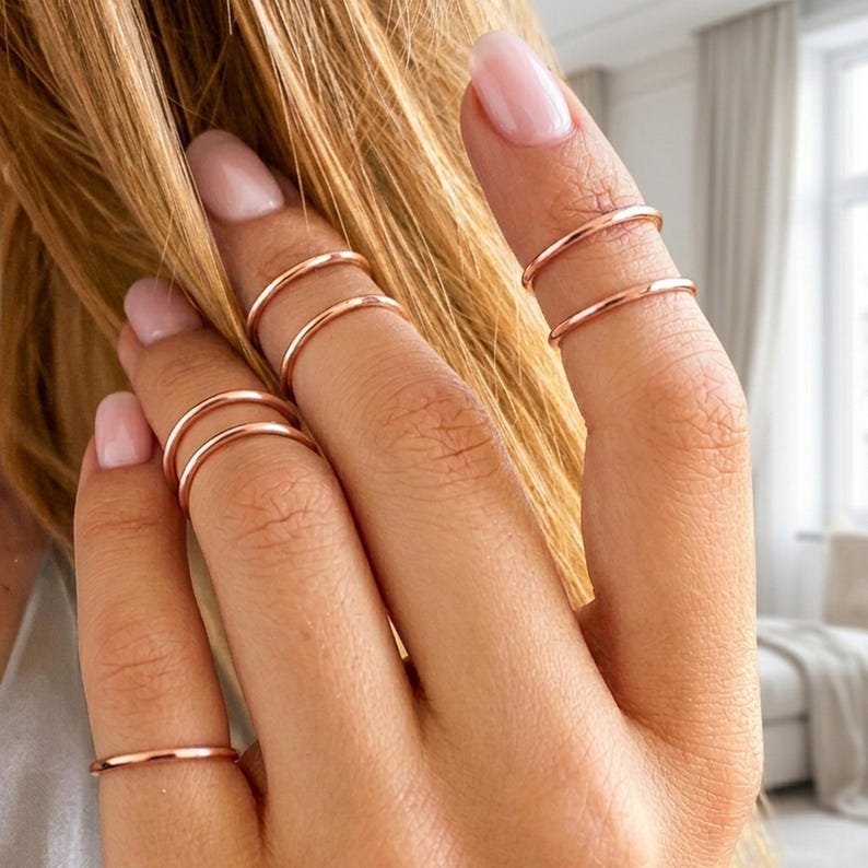 May include: A hand wearing multiple thin, rose gold rings on several fingers. The rings are simple bands, and the skin tone is fair. The background is blurred, suggesting a focus on the jewelry.
