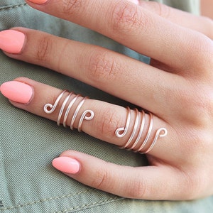 May include: A rose gold wire ring with multiple spirals. The ring is worn on the middle finger of a hand with pink nail polish.