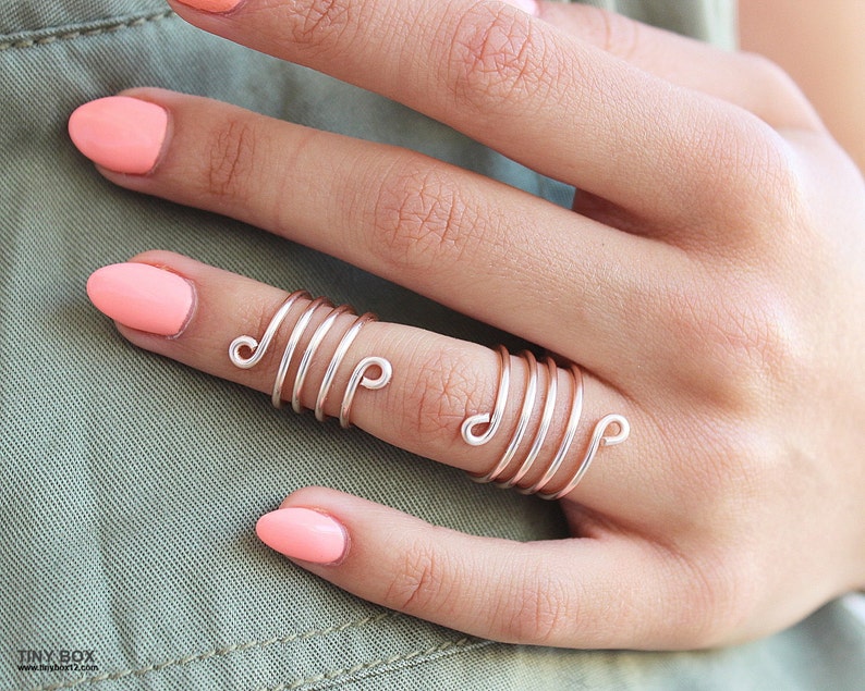 May include: A rose gold wire ring with multiple spirals. The ring is worn on the middle finger of a hand with pink nail polish.