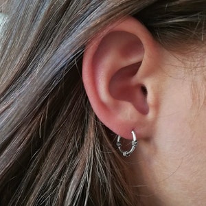 May include: A silver hoop earring with a textured design. The earring is worn in the left earlobe.