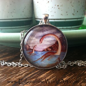 May include: A silver chain necklace with a round pendant featuring a watercolor painting of two plesiosaurs in a blue body of water. The pendant is set in a silver bezel and has a cloudy sky background.