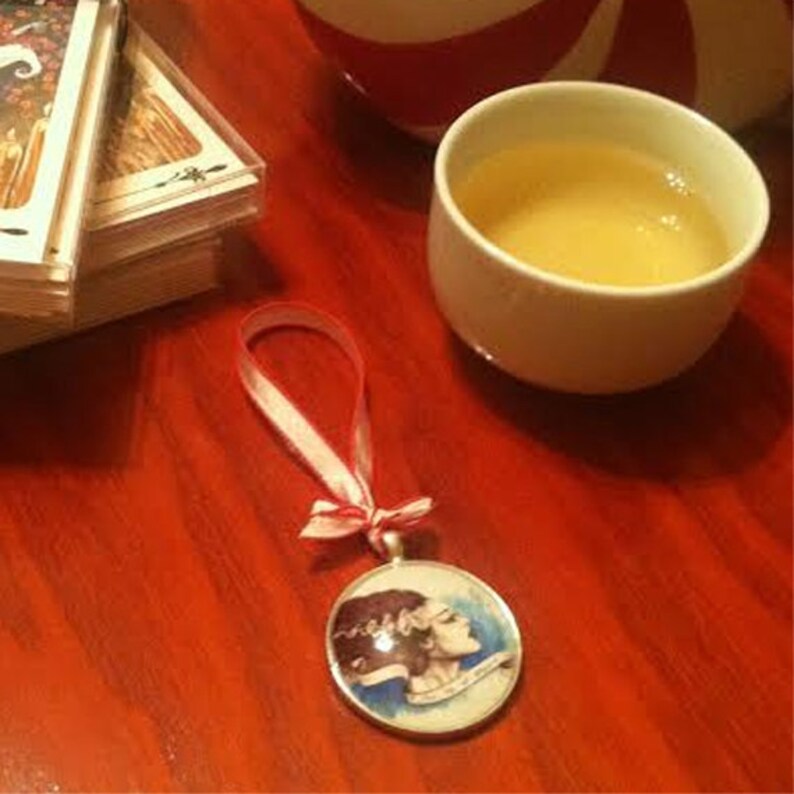 May include: A white ceramic teacup with a red and white striped teacup on the table. A white ribbon with red trim is tied around a round ornament with a drawing of a woman's face.