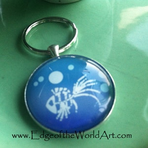 May include: A silver key chain with a blue glass charm featuring a white silhouette of a fish with bubbles. The charm has a gradient of blue colors. The text "www.EdgeoftheWorldArt.com" is printed below the charm.