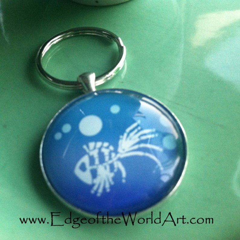 May include: A silver key chain with a blue glass charm featuring a white silhouette of a fish with bubbles. The charm has a gradient of blue colors. The text "www.EdgeoftheWorldArt.com" is printed below the charm.
