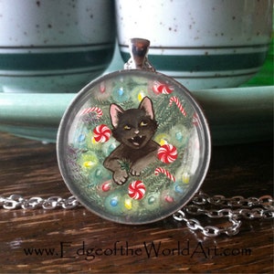 May include: A black cat with green eyes peeks out from a Christmas wreath with red and white candy canes, green pine branches, and colorful lights. The wreath is inside a clear glass pendant on a silver chain.