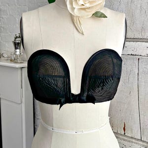 May include: Black strapless bra with a sheer overlay, displayed on a mannequin. The bra features a deep V-neckline and a small bow detail. A white ribbon is wrapped around the mannequin's waist. A large cream-colored rose adorns the mannequin's shoulder.