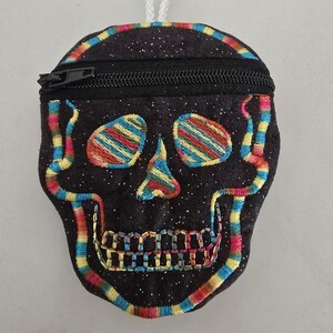 May include: A black skull-shaped coin purse with a zipper closure and a key ring. The skull design is embroidered with colorful stripes of red, yellow, and blue. The purse has a glittery texture.