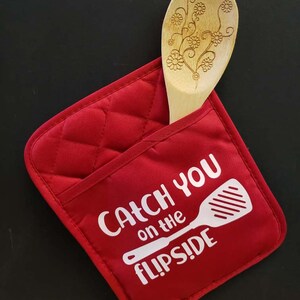 May include: A red quilted pot holder with the text "Catch You on the Flip Side" in white, holding a wooden spoon with a floral design. The pot holder is a kitchen accessory.