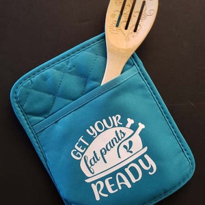May include: Teal quilted oven mitt with the text "GET YOUR fat pants READY" in white. A wooden slotted spoon is in the mitt's pocket. The mitt is on a black surface.