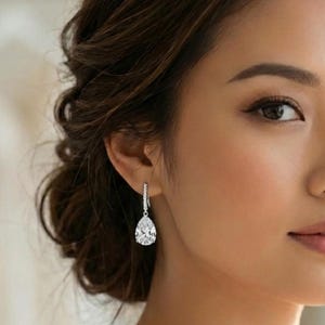 May include: A silver-toned drop earring featuring a single pear-shaped clear gemstone suspended from a small, delicate hook. The earring is crafted with a polished metal setting that holds the faceted stone, creating a bright, reflective appearance.