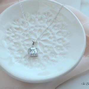 May include: A silver necklace with a square-shaped pendant featuring a large clear cubic zirconia stone surrounded by smaller stones. The necklace is displayed on a white ceramic dish with a decorative design.