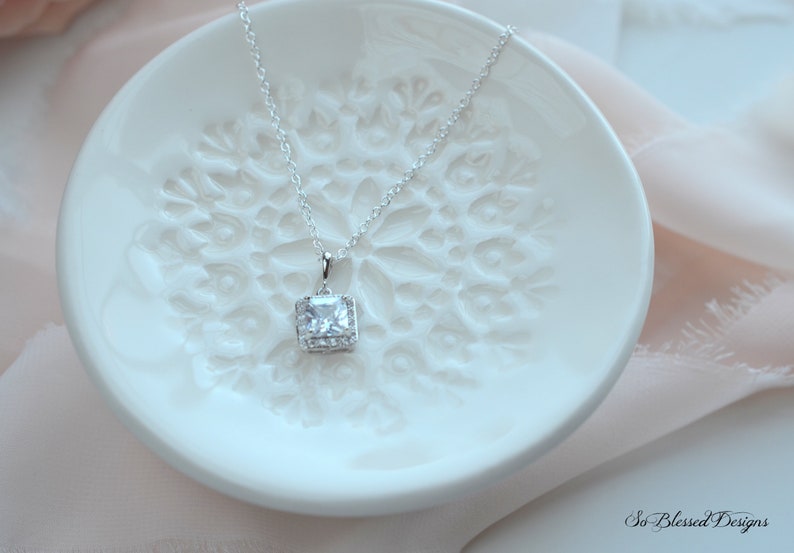 May include: A silver necklace with a square-shaped pendant featuring a large clear cubic zirconia stone surrounded by smaller stones. The necklace is displayed on a white ceramic dish with a decorative design.