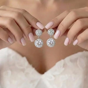 May include: Elegant silver earrings with a sparkling, round, clear gemstone surrounded by smaller stones. The earrings are held up by a person with manicured nails, against a white background.