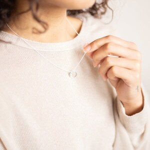 Pu&ograve; includere: Una collana d'argento con un semplice ciondolo a forma di cuore. La collana &egrave; indossata da una persona con i capelli ricci.