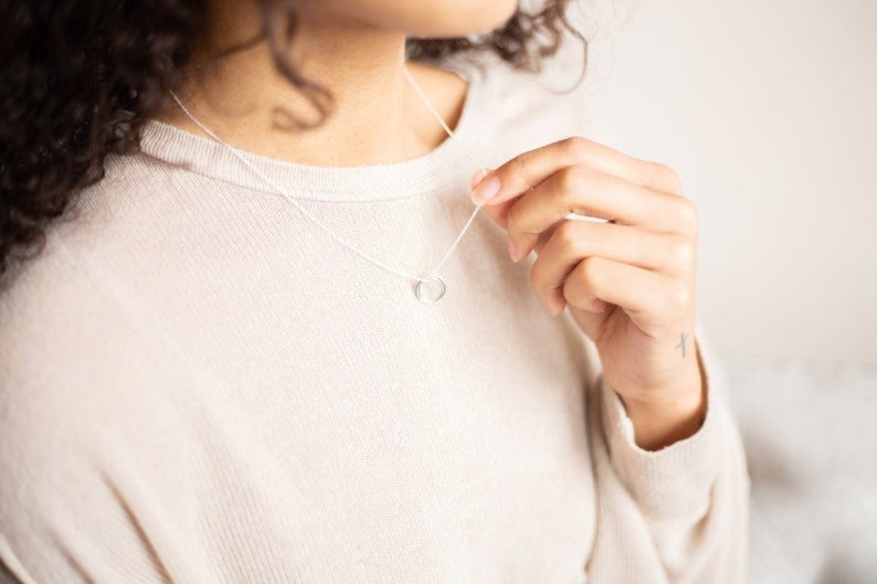 Pu&ograve; includere: Una collana d'argento con un semplice ciondolo a forma di cuore. La collana &egrave; indossata da una persona con i capelli ricci.