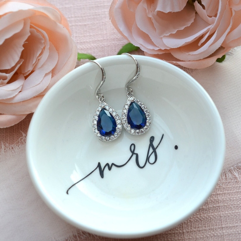 May include: A pair of teardrop-shaped earrings with a blue sapphire stone surrounded by a halo of smaller clear stones. The earrings are on a white dish with the word "mrs." written in black.