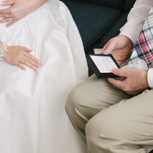 Puede incluir: Una persona con un vestido blanco se sienta junto a una persona que lleva una camisa blanca y una corbata a cuadros. La persona con la camisa blanca sostiene una peque&ntilde;a caja negra con una tarjeta blanca en su interior.