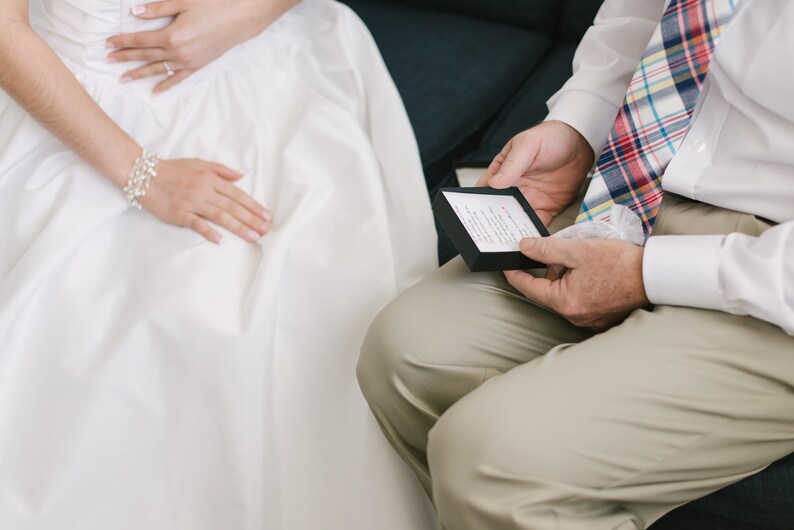 Puede incluir: Una persona con un vestido blanco se sienta junto a una persona que lleva una camisa blanca y una corbata a cuadros. La persona con la camisa blanca sostiene una peque&ntilde;a caja negra con una tarjeta blanca en su interior.