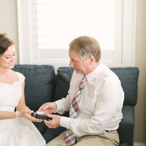 Puede incluir: Una novia con un vestido blanco recibe un regalo de un hombre con una camisa blanca y una corbata de cuadros rojos y azules. Est&aacute;n sentados en un sof&aacute;.