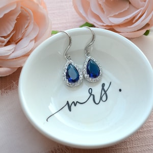May include: A pair of teardrop-shaped earrings with a blue gemstone surrounded by smaller clear stones. The earrings are on a white ceramic dish with the word "mrs." written in black.