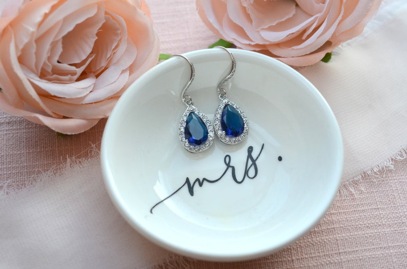 May include: A pair of teardrop-shaped earrings with a blue gemstone surrounded by smaller clear stones. The earrings are on a white ceramic dish with the word "mrs." written in black.
