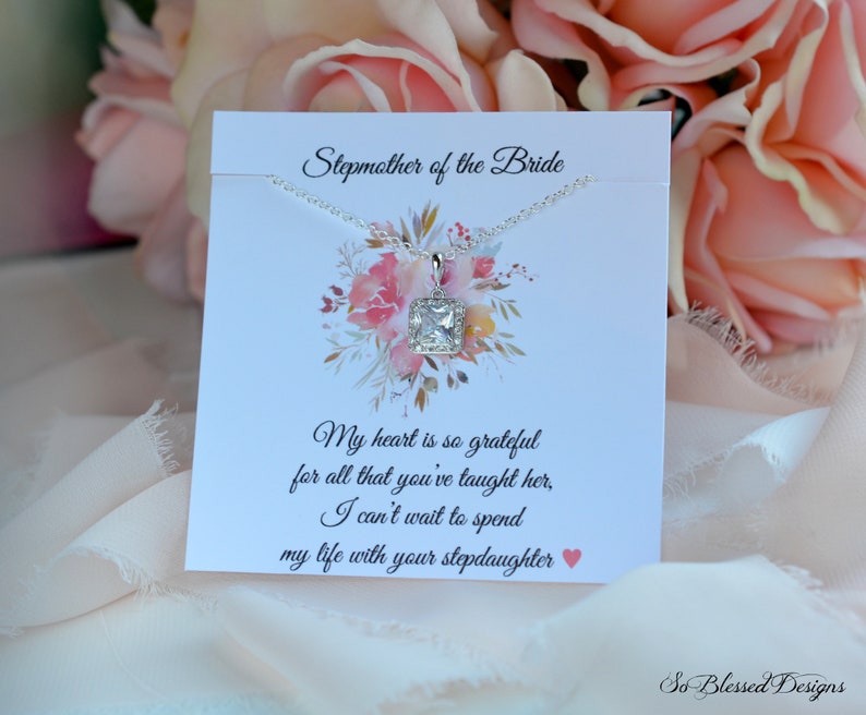 May include: A silver necklace with a square clear crystal pendant on a white card with the text "Stepmother of the Bride" and a message about being grateful for all that the stepmother has taught the bride. The card also includes the text "So Blessed Designs".