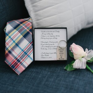 Puede incluir: Una peque&ntilde;a caja blanca con una corbata a cuadros en su interior, un llavero plateado con el texto "Always your little girl Rachel" y una rosa rosa con una cinta blanca.