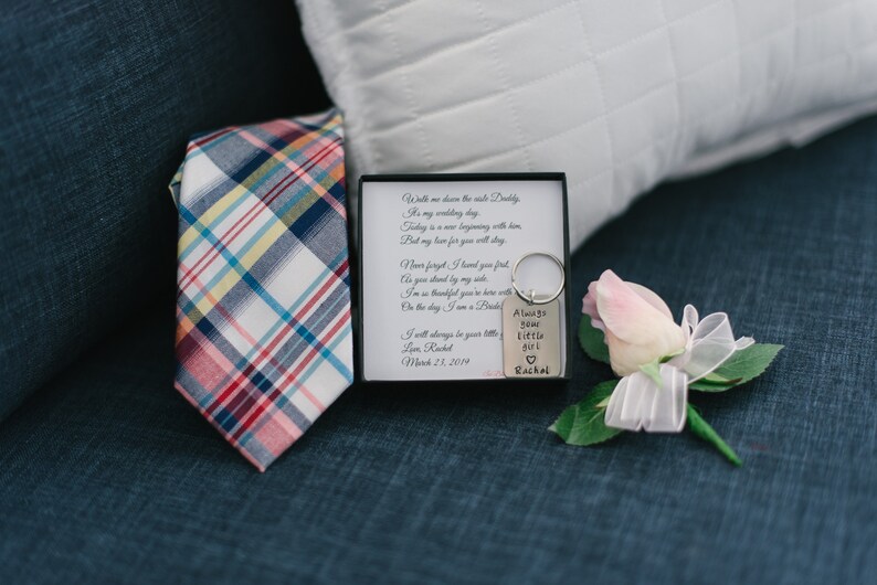 Puede incluir: Una peque&ntilde;a caja blanca con una corbata a cuadros en su interior, un llavero plateado con el texto "Always your little girl Rachel" y una rosa rosa con una cinta blanca.
