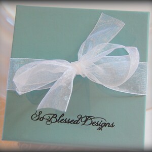 Pu&ograve; includere: Una scatola regalo blu chiaro con un nastro bianco trasparente legato in un fiocco. La scatola ha il testo "So Blessed Designs" stampato sul davanti.