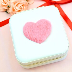 May include: A white square-shaped jewelry box with a zipper closure. The box has a pink fuzzy heart on the front.