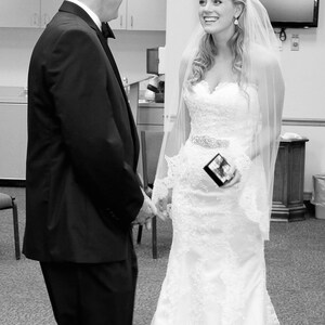 Puede incluir: Una novia con un vestido de novia blanco de encaje y velo sonr&iacute;e al novio, que lleva un traje negro y corbata.