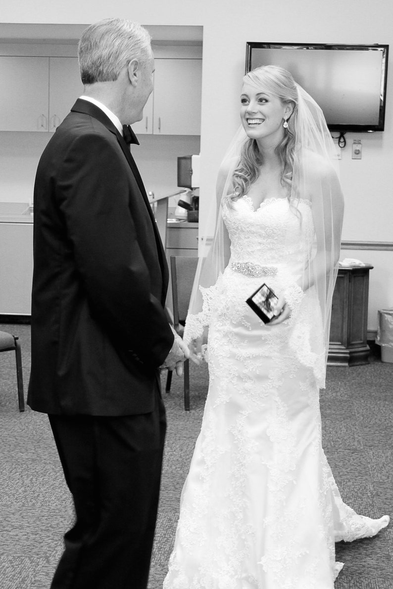 Puede incluir: Una novia con un vestido de novia blanco de encaje y velo sonr&iacute;e al novio, que lleva un traje negro y corbata.