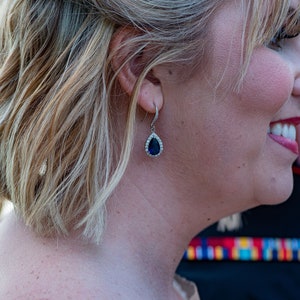 May include: A woman with blonde hair wearing a teardrop-shaped earring with a dark blue stone and a silver setting.