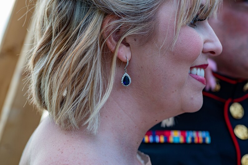 May include: A woman with blonde hair wearing a teardrop-shaped earring with a dark blue stone and a silver setting.