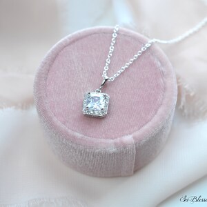 May include: A silver necklace with a square-shaped pendant featuring a large clear cubic zirconia stone surrounded by smaller cubic zirconia stones. The necklace is displayed in a pink velvet box.