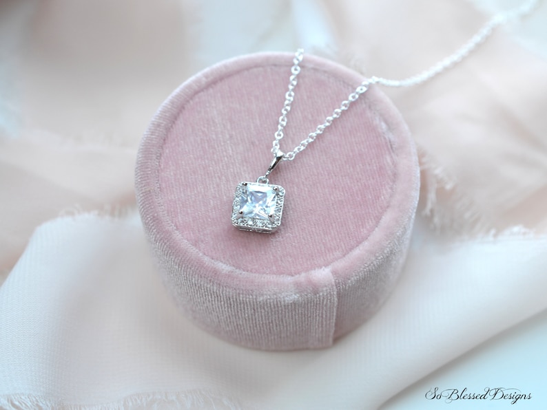 May include: A silver necklace with a square-shaped pendant featuring a large clear cubic zirconia stone surrounded by smaller cubic zirconia stones. The necklace is displayed in a pink velvet box.