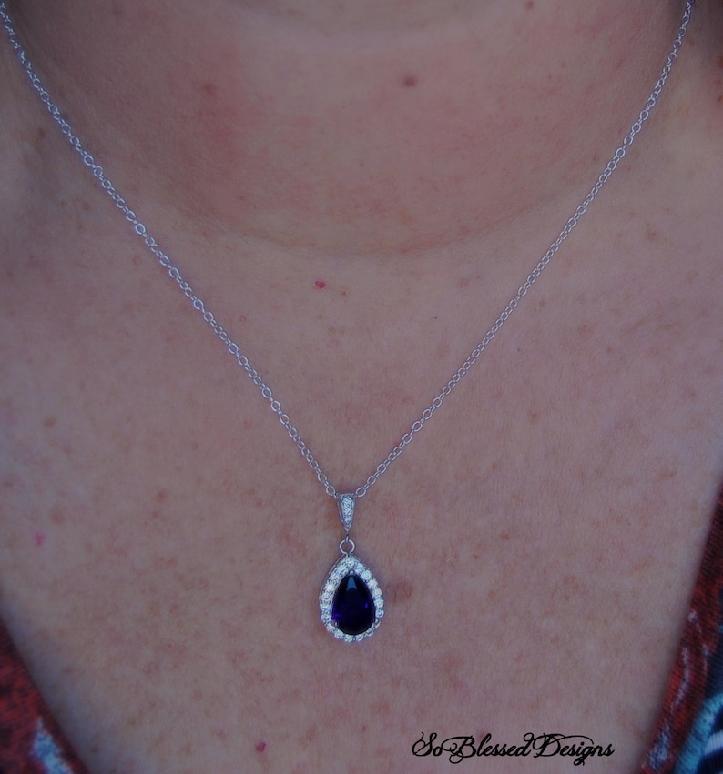 May include: A silver chain necklace with a teardrop-shaped pendant featuring a large, dark purple gemstone surrounded by smaller clear gemstones.