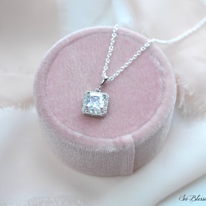 May include: A silver necklace with a square-shaped pendant featuring a large clear cubic zirconia stone surrounded by smaller cubic zirconia stones. The necklace is displayed in a pink velvet box.