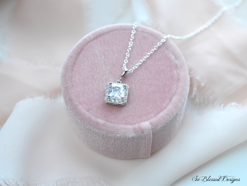 May include: A silver necklace with a square-shaped pendant featuring a large clear cubic zirconia stone surrounded by smaller cubic zirconia stones. The necklace is displayed in a pink velvet box.