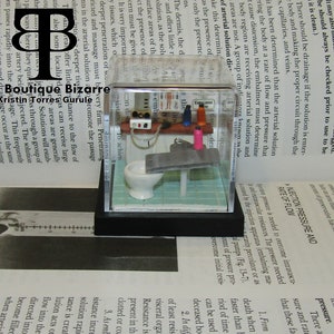 May include: A miniature bathroom scene in a clear acrylic box. The bathroom features a white toilet, a small sink, a towel rack, and a small shelf with various miniature toiletries. The box is sitting on a book with the title "The Boutique Bizarre" visible.