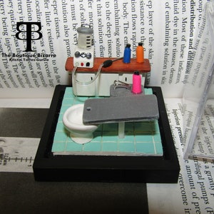 May include: A miniature medical scene with a white porcelain sink, a gray examination table, and a white medical machine with a clear tube. The scene is set on a blue and white tiled floor and framed in black.