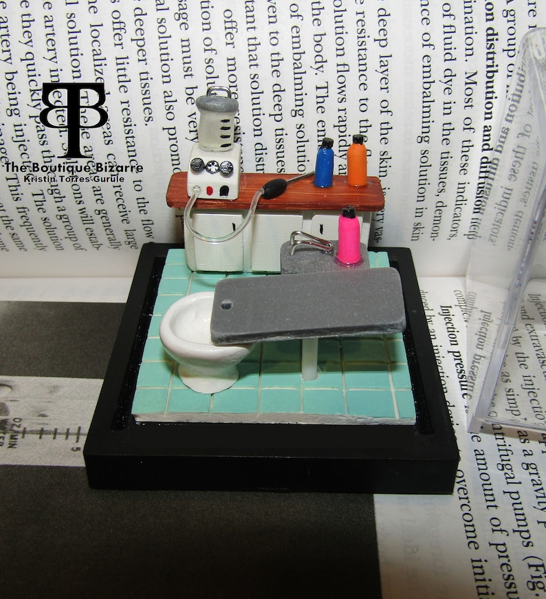 May include: A miniature medical scene with a white porcelain sink, a gray examination table, and a white medical machine with a clear tube. The scene is set on a blue and white tiled floor and framed in black.