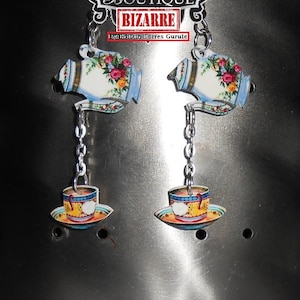 May include: A pair of earrings featuring a teapot and teacup design. The teapot is white with blue and gold accents and floral details. The teacup is yellow with blue and red accents. The earrings are attached to silver chains.
